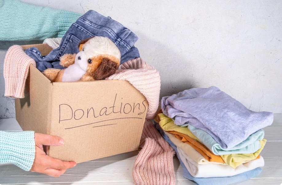 A woman packed up a cardboard box with gently used clothing items and a stuffed teddy bear. Next to the box are folded up clothes.